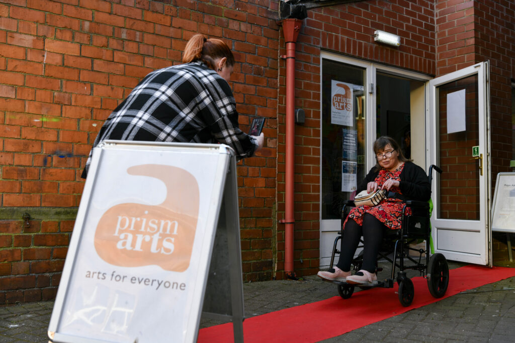A glamorous, female wheelchair user posing on a red carpet outside of Prism Arts.