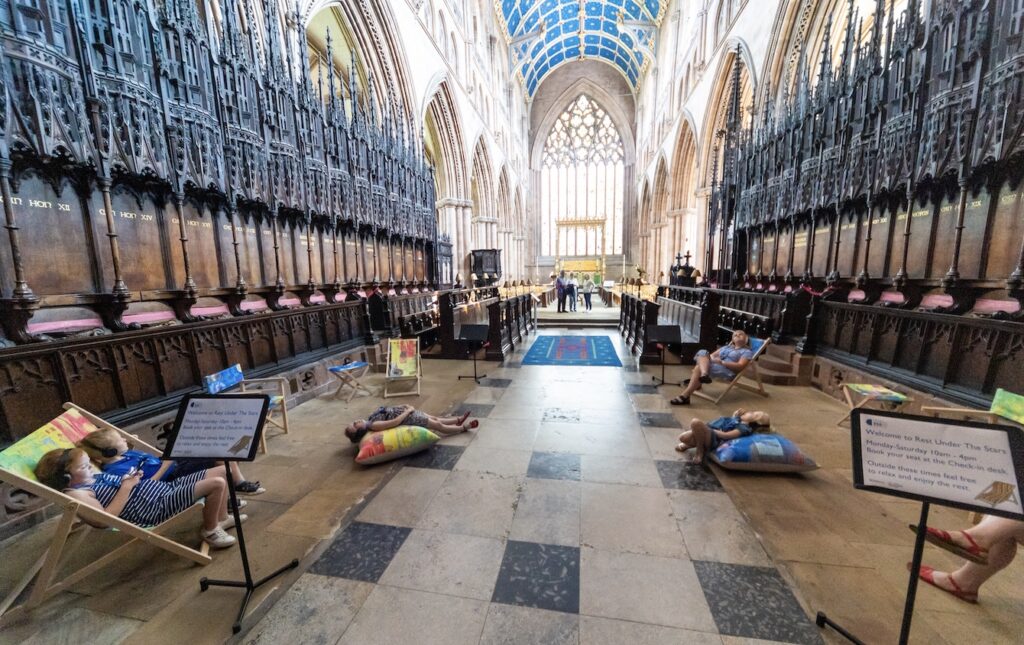 In a wide shot of Carlisle Cathedral, we see a group of people resting on colourful seating, looking up at the ceiling.