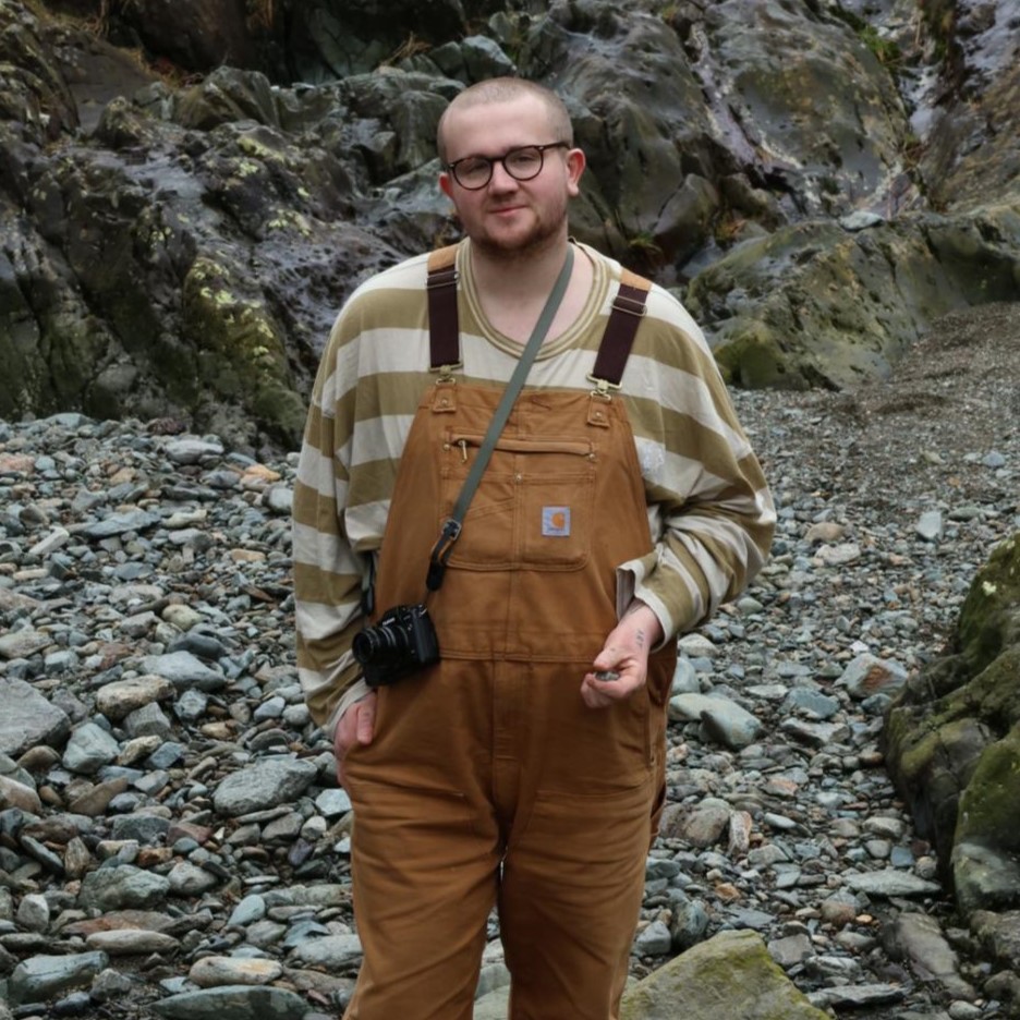 white person with brown dungarees and beige striped top, smiling calmly in a craggy countryside location. This person is non-binary and has black rimmed glasses.