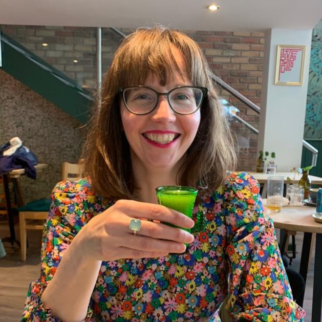 white woman in flower dress drinking small beverage in cafe. brown hair with black rimmed glasses. bright, happy smile.